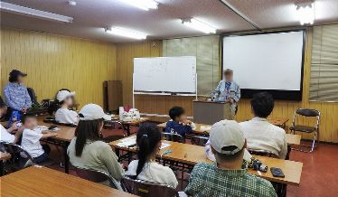 室内で先生がホワイトボードの前でセミの生態について説明しており、参加した親子連れが熱心に話を聞いている様子